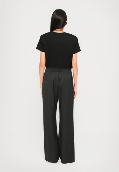 Woman with long dark hair wearing a black short-sleeve top and high-waisted loose black pants, standing facing away against a light background.