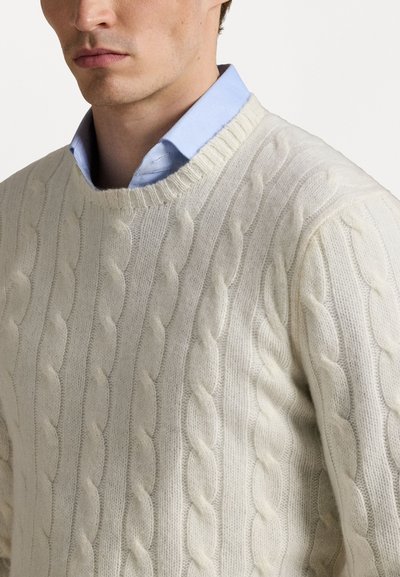 Pull en tricot torsadé de couleur crème, avec un col rond, des poignets côtelés et une texture douce, porté sur une chemise col bleu clair.