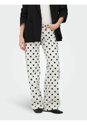 Personne portant un pantalon blanc à jambes larges avec des pois noirs, un blazer noir, un haut clair et des chaussures noires pointues, debout avec une main dans la poche.