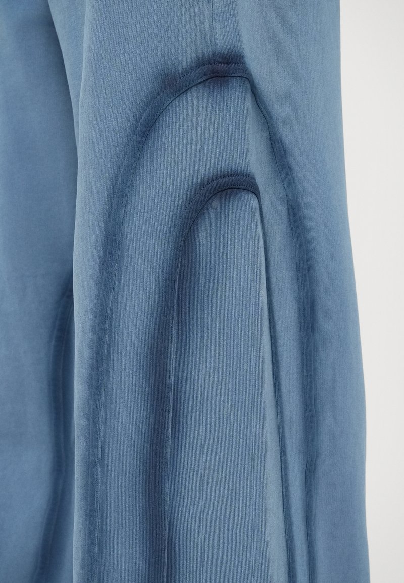 Närbild av bl�åa tygbyxor som visar en strukturerad, krökt sömdesign på benet mot en ljus bakgrund.