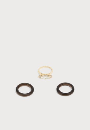 Anello d'oro con una grande pietra preziosa trasparente, posizionato sopra due anelli circolari marrone scuro su uno sfondo grigio chiaro.