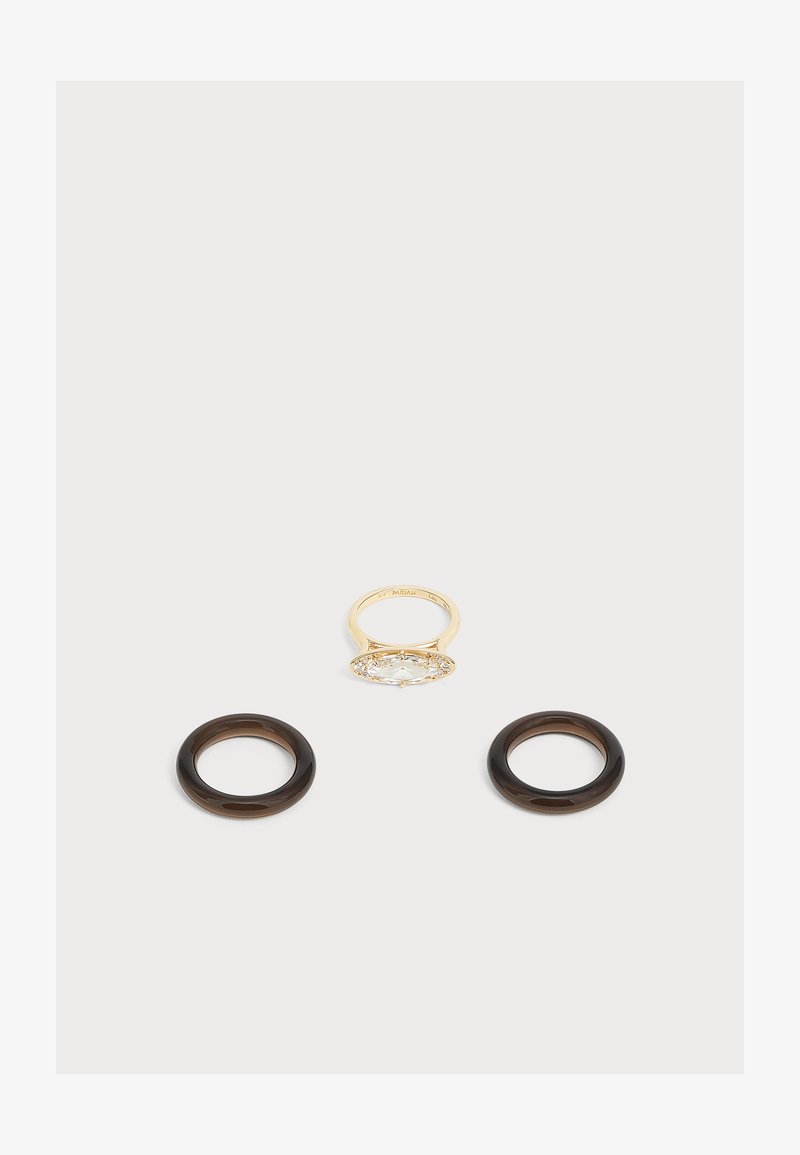 Bague en or avec une grande pierre précieuse claire, posée au-dessus de deux anneaux circulaires brun foncé sur un fond gris clair.