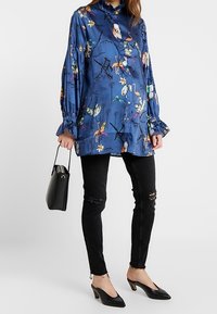 Blusa de satén azul con estampado colorido de flores y pájaros, que presenta un cuello alto y mangas fruncidas, combinada con jeans skinny negros rasgados y tacones negros.