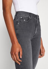 Jeans de mezclilla gris con corte ajustado. Presentan un logo en el bolsillo frontal, herrajes plateados y costuras mínimas. Textura suave.