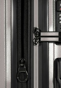 Primo piano delle cerniere argento e nere di una valigia, comprese due etichettate "REDOLZ," con guscio rigido testurizzato e serratura a combinazione integrata.