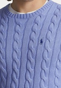 Ljuslila kabelstickad tröja med en liten mörkblå broderad hästlogotyp på bröstet, bärs över en vit crewneck-tröja.