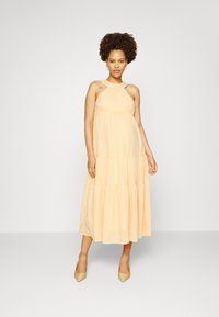 Vestido maxi em pêssego claro com decote halter, design de camadas de babados e um tecido texturizado. Usado com sapatos de bico fino bege.