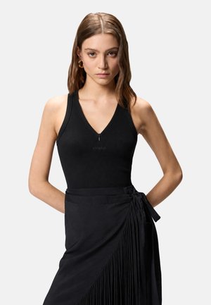 Giovane donna con capelli castano chiaro indossa un top nero senza maniche con scollo a V e una gonna nera a portafoglio con pieghe e legatura laterale.