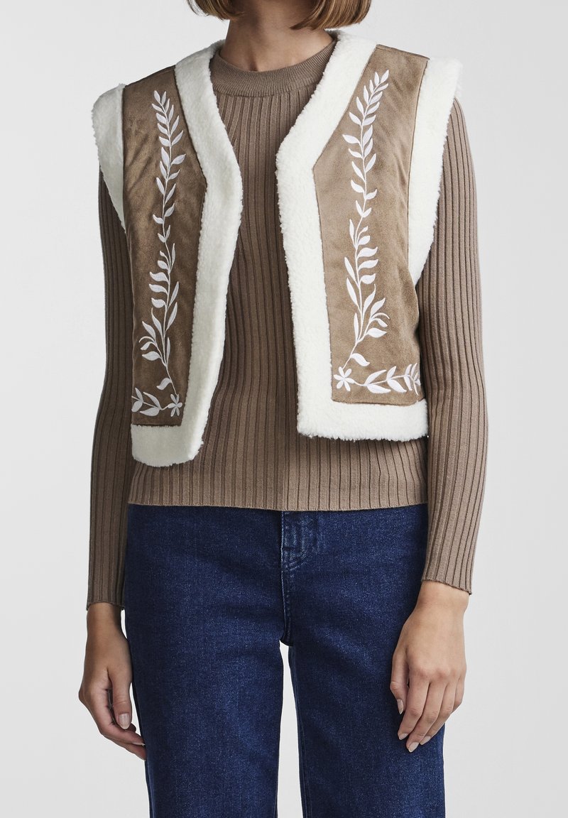 Brun konstläderväst med vit fleecekant, dekorerad med vita broderade bladmönster. Bärs över en ribbad brun topp och blå jeans.