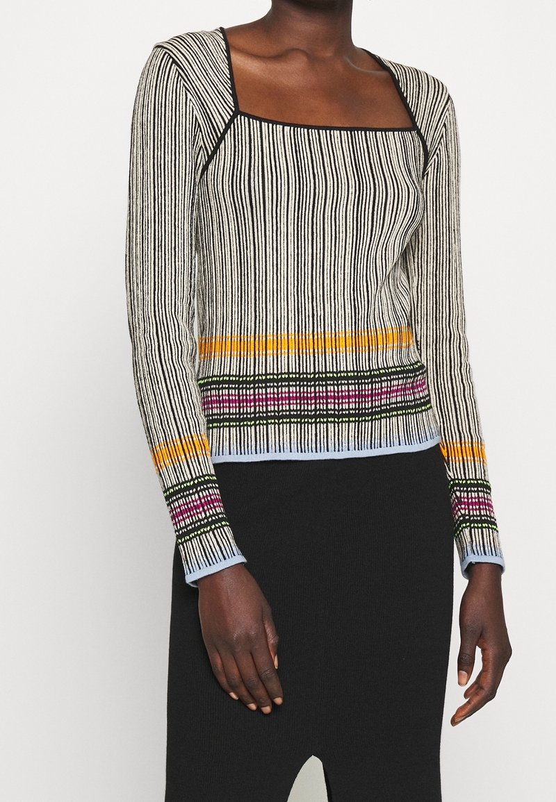 Personne à la peau noire portant une chemise à manches longues et col carré avec des rayures verticales noires et blanches et des motifs colorés près de l'ourlet et des poignets.