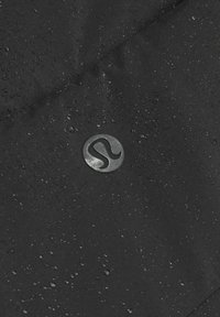 Melnā ūdensnecaurlaidīgā auduma ar matētu faktūru, kurā redzami mazi pilieni un apļveida logo iepresēts gaišākā nokrāsā.