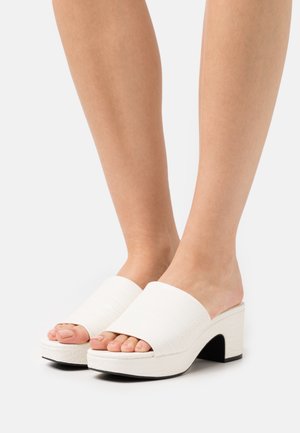 Mules blanches à talons avec un motif texturé en crocodile. Design ouvert à l'avant et larges brides, avec une semelle noire pour le contraste.