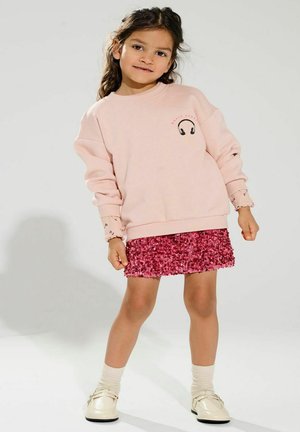 Lyserød sweatshirts med hovedtelefon grafik, parret med en glitrende fuchsia nederdel. Model iført cremefarvede sokker og skinnende sko.