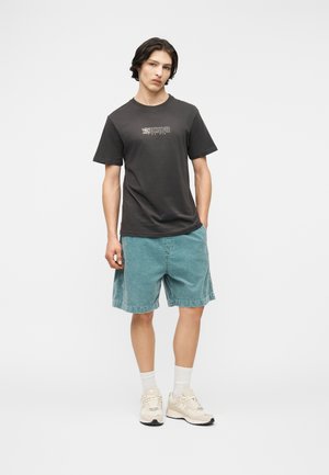 Νέος άνδρας που στέκεται με το χέρι στην τσέπη, φορώντας μαύρο t-shirt με σχέδιο, πετρόλ σορτς, άσπρες κάλτσες και μπεζ αθλητικά παπούτσια σε λευκό φόντο.