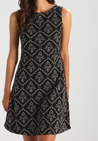 Svart ärmlös klänning med ett texturerat damaskmönster i kontrasterande beige och guld, med rund halsringning och utsvängd silhuett.