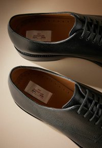 Sorte lædersko med tekstureret overflade, rund tå og snørebåndsdesign. Har en tan inderside og en mærkat, der står "Next Signature Italian Leather."