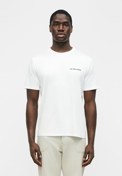 Άνδρας που φοράει απλό λευκό κοντομάνικο μπλουζάκι με το κείμενο "LES BENJAMINS" στο αριστερό μέρος του στήθους και ανοιχτόχρωμο παντελόνι, στέκεται μπροστά σε λευκό φόντο.