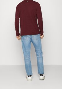 Langärmliges weinrotes Hemd, blaue Jeans und weiße Turnschuhe mit schwarzen Akzenten. Das Hemd hat einen runden Halsausschnitt und eng anliegende Ärmel.