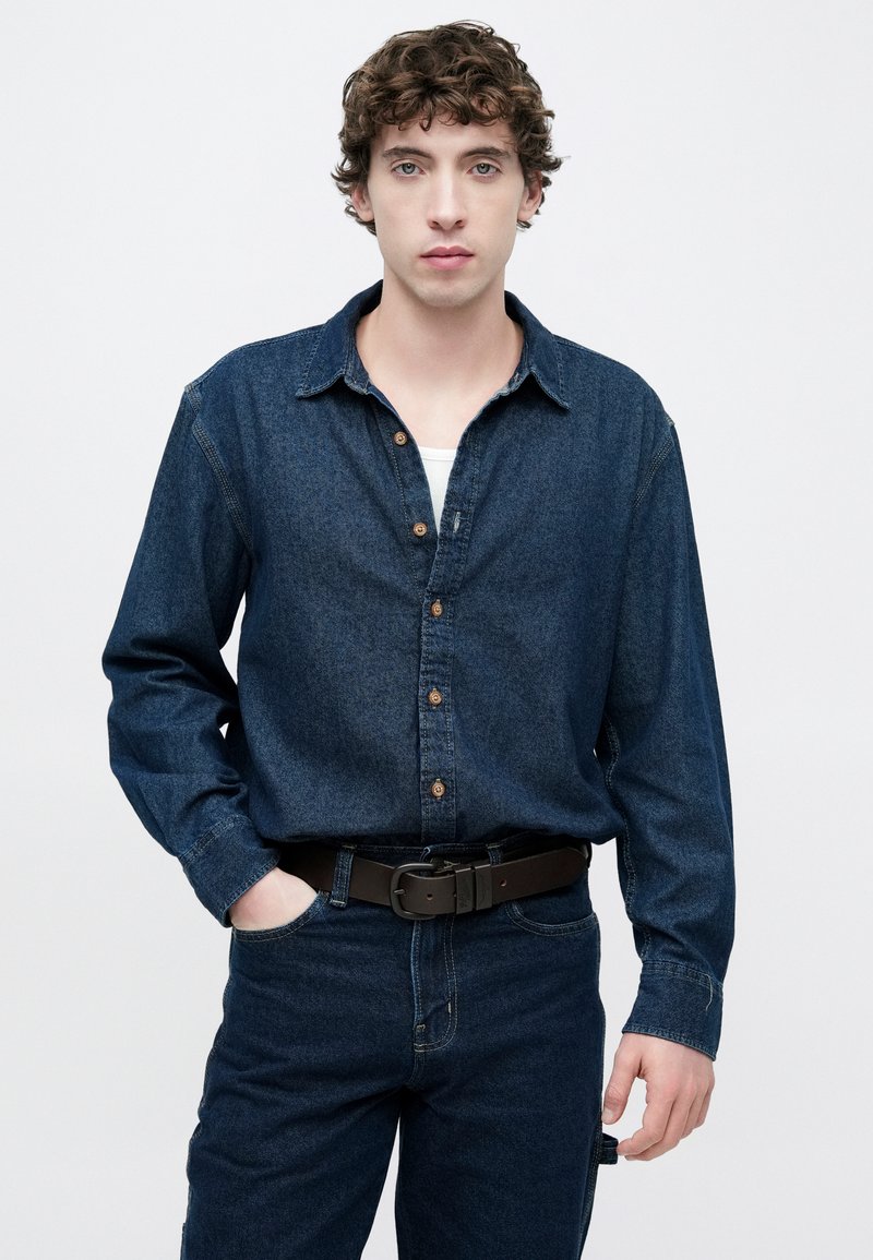 Denim shirt with long sleeves, dark blue color, button-down front, and a casual fit. Paired with dark blue jeans and a black belt.