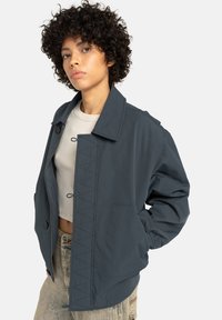 Young person with curly hair wearing dark gray buttoned jacket over light crop top and beige pants, standing with one hand in pocket.