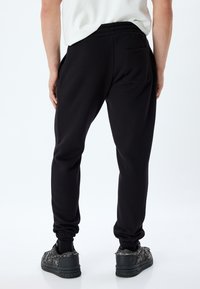 Svarta joggingbyxor med elastisk midja, avsmalnande ben och en bakficka. Materialet verkar mjukt med en slät textur.