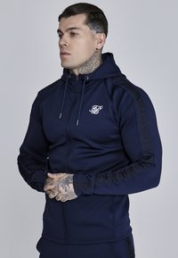 Mornarska dukserica na zip, izrađena od rastezljivog materijala, s logom na prsima i crnim detaljima duž rukava.