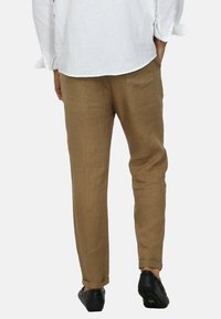 Pantaloni in lino marrone con vestibilità affusolata, dotati di tasche laterali e orli arrotolati, abbinati a una camicia bianca a maniche lunghe e scarpe nere.