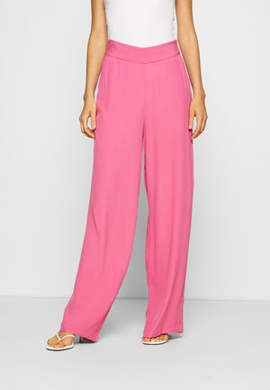 Mujer con pantalones rosa de talle alto y piernas anchas con una pretina en forma de V y sandalias blancas de tacón sobre un fondo liso.