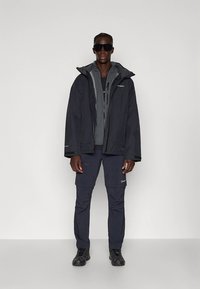 Tume veekindel jope kapuutsiga, kantud tumeda halli särgi peal. Tumedad püksid taskutega ja mustad kingad. Modell kannab tumedaid päikeseprille.