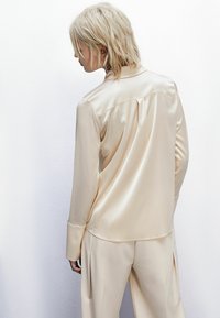 Camicia in satin crema con maniche lunghe, colletto con bottoni e dettagli plissettati sul retro, abbinata a pantaloni a vita alta coordinati.