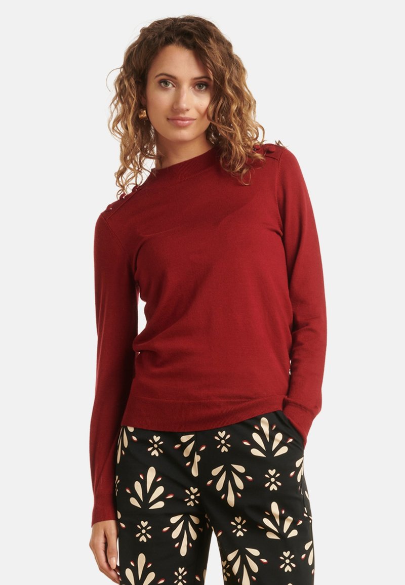 Red long-sleeve sweater with a high neckline and shoulder detailing. Worn with black patterned pants featuring cream and red floral motifs.