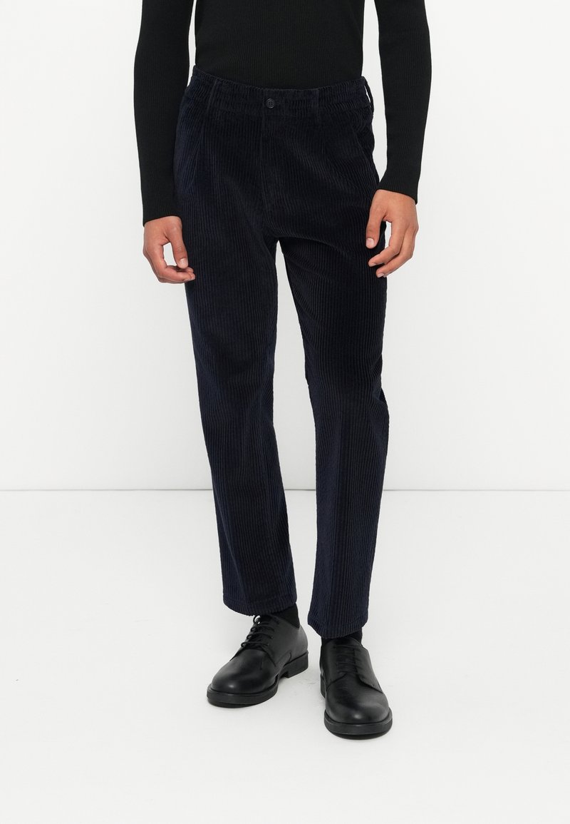 Personne portant un pull noir côtelé, un pantalon en velours bleu foncé et des chaussures habillées noires, debout devant un fond blanc uni.