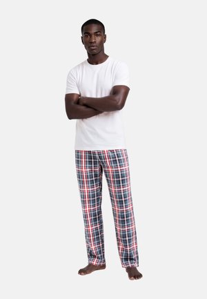 Man staat op blote voeten met gekruiste armen en draagt een effen wit T-shirt en rood, wit en blauw geruite pyjamabroek tegen een witte achtergrond.