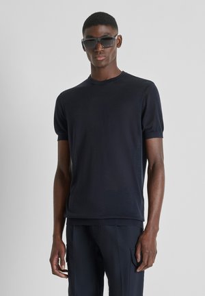 Mladić u tamnoj majici kratkih rukava, tamnim hlačama s džepovima i velikim crnim sunčanim naočalama, stoji na jednostavnoj svijetloj pozadini.