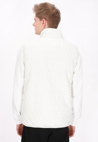 Weiße gesteppte Weste mit hochgeschlossenem Kragen, texturiertem Oberflächen und leichtem Design. Getragen über einem Langarmshirt.
