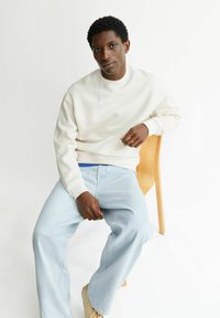 Felpa beige chiaro con scollo rotondo, abbinata a jeans ampi color azzurro chiaro. Seduta su una sedia di legno davanti a uno sfondo bianco.