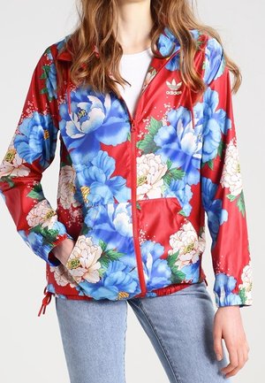 Woman wearing red Adidas zip-up jacket with large blue and white floral pattern and light blue jeans, hands partially in pockets.
