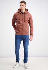 Brūns džemperis ar rāvējslēdzēju un kapuci ar auklu, ar ribotām aprocēm un apakšmalu, kopā ar ziliem džinsiem un baltiem sporta apaviem. Audums izskatās mīksts.