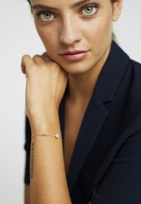 Pulsera de oro con un pequeño charm circular. Cadena suave y delicada. El brazo del modelo lleva un tatuaje. Chaqueta de tela oscura de fondo.