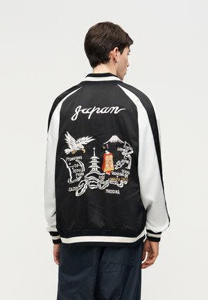 Hombre con chaqueta blanca y negra que lleva bordados en la parte trasera un mapa de Japón, un águila, una pagoda, una geisha y nombres de ciudades.