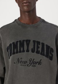 Donkergrijze sweatshirt met ronde hals. Heeft een gestructureerde zwarte borduurtekst van "TOMMY JEANS" en een scriptdetail "New York sinds 1985".