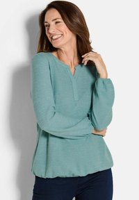 Lächelnde Frau mit braunen Haaren, die eine langärmelige türkisfarbene Bluse und dunkle Hosen trägt, vor einem weißen Hintergrund steht.