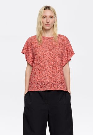 Top rosso a fantasia con maniche corte e vestibilità morbida. Il tessuto è leggero con una sottile texture. Abbinato a pantaloni neri larghi.
