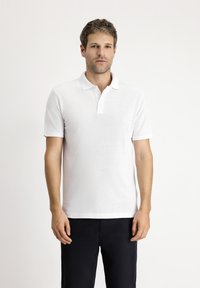 Biała koszulka polo z teksturowanej tkaniny, z kołnierzykiem i trzema guzikami. Noszona z ciemnymi spodniami, model stoi na jasnym tle.