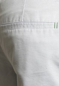 Tissu gris clair avec une finition texturée, présentant un détail de couture horizontale et un subtil accent vert, typiquement utilisé dans la conception de pantalons.