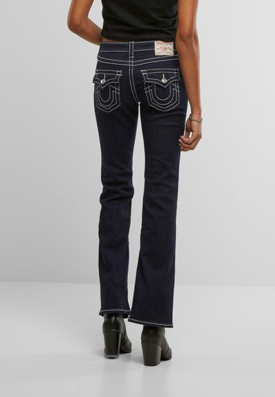 Jean évasé bleu foncé avec des coutures blanches contrastantes, présentant des poches arrière distinctives et une étiquette True Religion sur la taille.