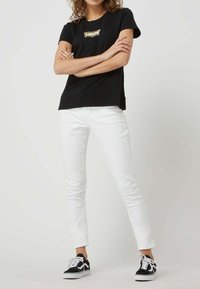 Camiseta de algodón negra de manga corta con un logo dorado. Combinada con jeans ajustados blancos y zapatillas negras con suelas y cordones blancos.