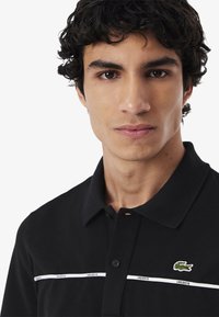 Musta polo-paita teksturoidusta kankaasta, jossa valkoinen raita ja "LACOSTE"-teksti sekä vihreä krokotiililogo rinnassa.