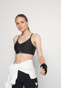 Schwarzer Sport-BH mit verstellbaren Trägern und Logo-Details. Glatte Textur und unterstützendes Design. Kombiniert mit einem weißen Sweatshirt, das an der Taille gebunden ist.