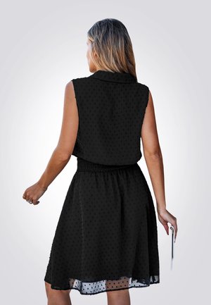 Een vrouw met lang, steil haar draagt een mouwloos zwart jurkje met een geraffineerde taille, terwijl ze een telefoon in haar rechterhand houdt.